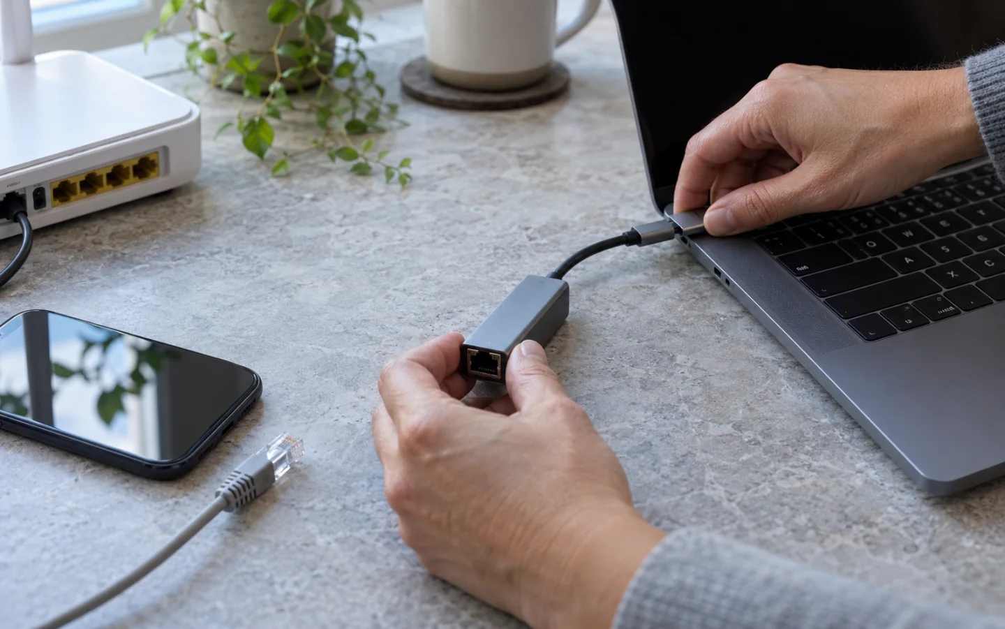 手部正在为笔记本连接USB-C网络转接器，旁边放有手机和家庭网关